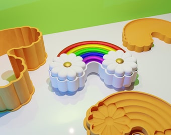 Op de afbeelding: Een regenboogvormige koekjesuitsteker met een madeliefjesontwerp aan elk uiteinde. De uitsteker is gemaakt van oranje plastic en wordt getoond op een wit oppervlak.
