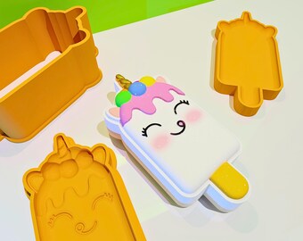 May include: A set of yellow plastic popsicle-shaped molds and a white and pink unicorn popsicle. The unicorn popsicle has a yellow stick, a pink top, and colorful sprinkles. The molds are in various shapes.