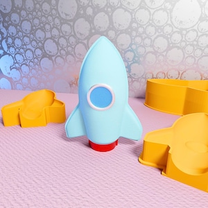 May include: A light blue rocket-shaped object with a red base and a blue circle in the center. Two yellow rocket-shaped cookie cutters are also present. The objects are on a pink surface with a silver and white patterned background.