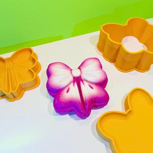 Puede incluir: Tres moldes de silicona amarillos en forma de mariposa, flor y lazo. El molde en forma de lazo está lleno de un pastel en forma de lazo rosa y blanco.