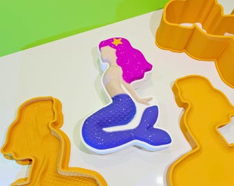 May include: A set of four silicone molds for baking mermaid-themed cakes or treats. The molds are yellow and feature a mermaid with a purple tail, a star on her head, and a mermaid with a long tail.