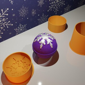 May include: A purple bath bomb with a silver snowflake design, alongside orange moulds. The background features a blue wall with white snowflake patterns. The scene is set on a white surface.