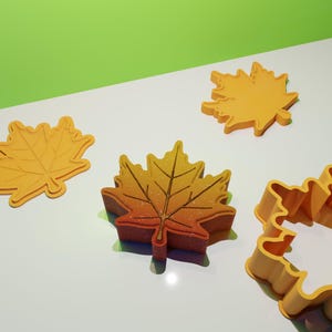 Könnte beinhalten: Vier verschiedene Blätter-förmige Ausstecher, zwei sind gelb und zwei sind orange. Die orangen Ausstecher haben ein detaillierteres Design mit Adern und einer strukturierten Oberfläche.