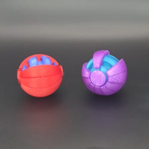 May include: Two spherical, 3D-printed puzzle toys. One is red with blue accents, the other is purple with blue accents. The toys have a segmented design, with interlocking pieces. The toys are designed to be taken apart and reassembled.