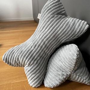 Könnte beinhalten: Ein graues Cordkissen in Knochenform. Das Kissen ist aus weichem, plüschigem Stoff und hat eine gerippte Textur.