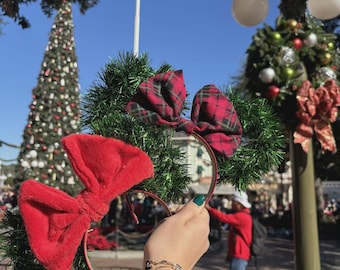 May include: Red and green Christmas-themed mouse ear headbands with plaid bows. The ears are decorated with green garland. A person is holding the headbands in front of a Christmas tree and a lamppost decorated with Christmas ornaments.