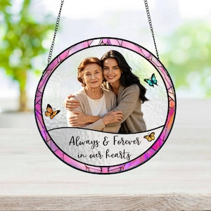 Puede incluir: Marco de fotos redondo de vidriera con una foto de dos mujeres abrazándose. El marco tiene un borde rosa y morado, mariposas y el texto "Always & Forever in our hearts". Cuelga de una cadena negra.
