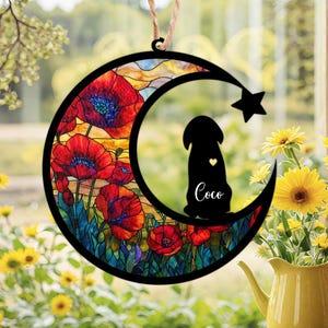 May include: A decorative hanging ornament featuring a black silhouette of a dog inside a crescent moon shape. The moon design includes red poppy flowers and a star. The dog's name, Coco, is written below. A jute string is attached for hanging.