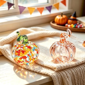 Puede incluir: Dos figuras de calabaza de cristal. La calabaza de la izquierda tiene un cuerpo transparente con manchas multicolores tipo confeti y un tallo verde. La calabaza de la derecha es transparente con patrones de remolinos naranjas y un tallo a juego.