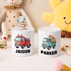 May include: Two white ceramic money banks with cartoon vehicle designs. One features a red fire truck with the name "JAISON" below. The other has a blue truck carrying boxes, with the name "PARKER". Both have money inserted.