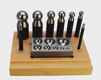 Könnte beinhalten: Ein Metall-Dapping-Block-Set bestehend aus einem rechteckigen Stahlblock mit mehreren halbkugelförmigen Vertiefungen und verschiedenen zylindrischen Stahlstempeln mit abgerundeten Spitzen, angeordnet auf einem hellen Holzsockel.