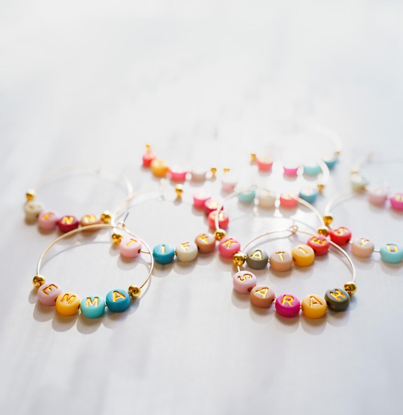 May include: Gold hoop earrings with colorful letter beads spelling out names. The beads are in various colors, including pink, blue, yellow, and green. The earrings are displayed on a white surface.
