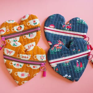 May include: Heart-shaped quilted pouches in mustard yellow and navy blue. The yellow pouches feature teacup and butterfly patterns, while the blue ones showcase mushroom and forest scenes. Each has a zipper and tassel.