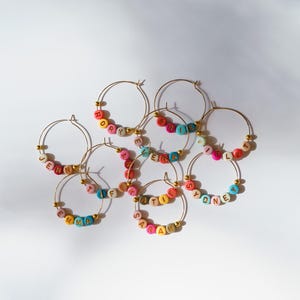 May include: Gold hoop earrings with colorful letter beads spelling out names like "Emma", "Sydney", and "Jenna". The beads are in various colors, including pink, blue, yellow, and orange. The earrings are displayed on a white surface.