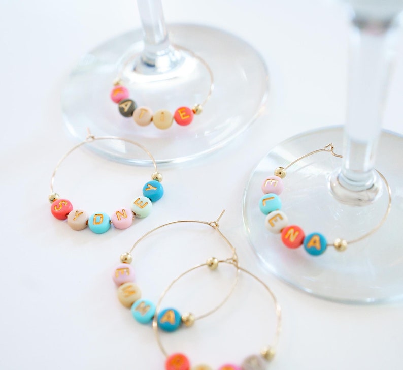 May include: Gold-colored hoop wine charms with colorful lettered beads spelling out names. The beads are in various colors, including pink, blue, yellow, and orange. The charms are attached to wine glasses.