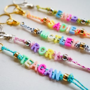 May include: Close-up of several colorful keychains with names spelled out in pastel letter beads. Each keychain has a different color cord, metal clasp, and decorative beads. Names include: EMILY, ADDIE, MOTLEY, RORY, and TEAGAN.