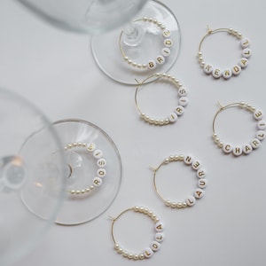 Könnte beinhalten: Goldene Weinringe mit weißen Perlen und Perlenakzenten. Jeder Ringbuchstabe buchstabiert einen Namen, darunter "Bride", "Iris", "Kendall", "Charlie", "Lauren" und "Lola". Die Ringe sind auf einer weißen Oberfläche angeordnet.