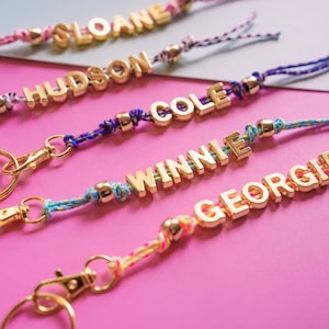 May include: Close-up of personalized keychains with gold-colored letter beads spelling out names: SLOANE, HUDSON, COLE, WINNIE, and GEORGIE. The keychains have colorful braided cords and gold-tone clasps, set against a pink and gray background.