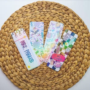 以下が含まれることがあります： 鉛筆、リンゴ、テキスト「MRS. PARKER」など、さまざまなデザインが施された、教師をテーマにした4つのカラフルなブックマーク。 ブックマークは、織られた円形の表面にあります。