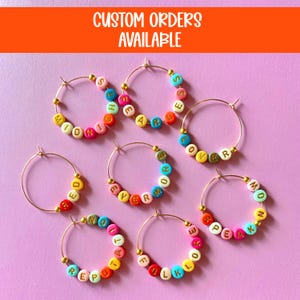 May include: Gold hoop earrings with colorful letter beads spelling out words like "MIDNIGHT", "SPEAK", and "FOREVER". The image also includes the text "CUSTOM ORDERS AVAILABLE" in white letters on an orange background.