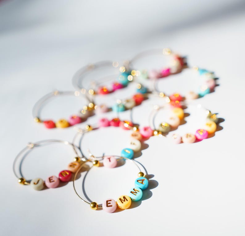 May include: Several hoop earrings with colorful beads spelling out names. The earrings have gold accents and are arranged on a white surface. The beads are in various colors, including pink, blue, yellow, and green.