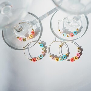May include: Four gold-colored wine glass charms with names spelled out in colorful beads. The names are Katie, Elena, Sydney, Emma, and Rory. The charms are on a white surface with the shadows of wine glasses.