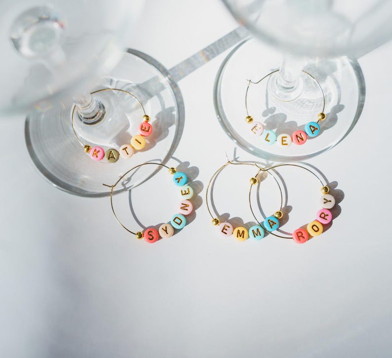 May include: Four gold-colored wine glass charms with names spelled out in colorful beads. The names are Katie, Elena, Sydney, Emma, and Rory. The charms are on a white surface with the shadows of wine glasses.