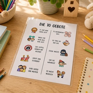 Puede incluir: Una hoja educativa plastificada titulada Die 10 Gebote que presenta diez mandamientos religiosos ilustrados en alemán con iconos coloridos, rodeada por una cesta de lápices de colores, un libro de historias bíblicas, un cuaderno y un peluche.