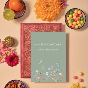 May include: A still life arrangement with a book titled "Mein Bibel-Lese-Tracker" and a pink patterned book. The image includes flowers, fruit, and snacks in small bowls, all set on a peach-coloured surface.