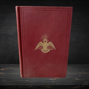Könnte beinhalten: Ein rotes Hardcover-Buch mit einem goldgeprägten Doppeladler-Emblem auf dem Vorderdeckel. Das Emblem zeigt eine Krone und ein strahlendes Dreieck über dem Adler sowie ein Banner mit Text unterhalb der Flügel des Adlers.