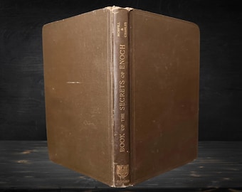 Pode incluir: Um livro antigo de capa dura marrom intitulado "Book of the Secrets of Enoch". A lombada exibe o título e o nome do autor em letras douradas. O livro está em pé, mostrando sua aparência envelhecida.