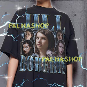 May include: Black t-shirt featuring a graphic of Julia Roberts with the text "PALMASHOP ROBERTS". The shirt has a crew neck and short sleeves. The design includes multiple images of Julia Roberts. The shirt is worn by a person.