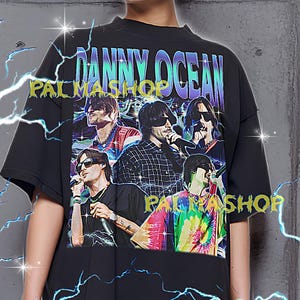 May include: Black oversized t-shirt featuring a graphic of a band with the text "DANNY OCEAN" and "PALMASHOP". The shirt has a crew neck and short sleeves. The model is wearing an XL shirt.