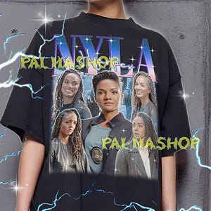 May include: Black oversized t-shirt featuring a graphic of several women and the text "NYLA PALMASHOP". The shirt has a crew neck and short sleeves. The text "PALMASHOP" is also printed on the shirt. The shirt is worn by a person.