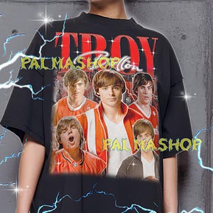 May include: Black oversized t-shirt featuring a graphic of five people in orange and white athletic wear. The word "TROY" is in red block letters above the graphic, with "PALMASHOP" in a stylized font below. The shirt has a vintage aesthetic.