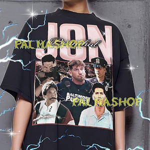 May include: Black t-shirt featuring a graphic of several men and the name "JON" in pink and white. The text "PALM Berthal" and "BALTIMORE PAL WASHOP" are also visible. The model is wearing an XL shirt.