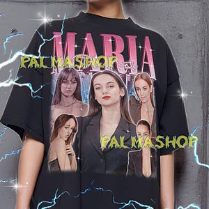 May include: Black oversized t-shirt featuring the name "MARIA" in pink, with images of women. The shirt also has the text "PALMASHOP" in green. The shirt is a unisex style.