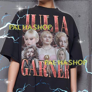 以下が含まれることがあります： ジュリア・ガーナーのグラフィックと「JULIA GARNER」の文字がメタリックゴールドでプリントされた黒いTシャツ。「PALMASHOP」の文字も入っています。モデルはXLサイズのシャツを着用。このユニセックスTシャツは通常サイズです。
