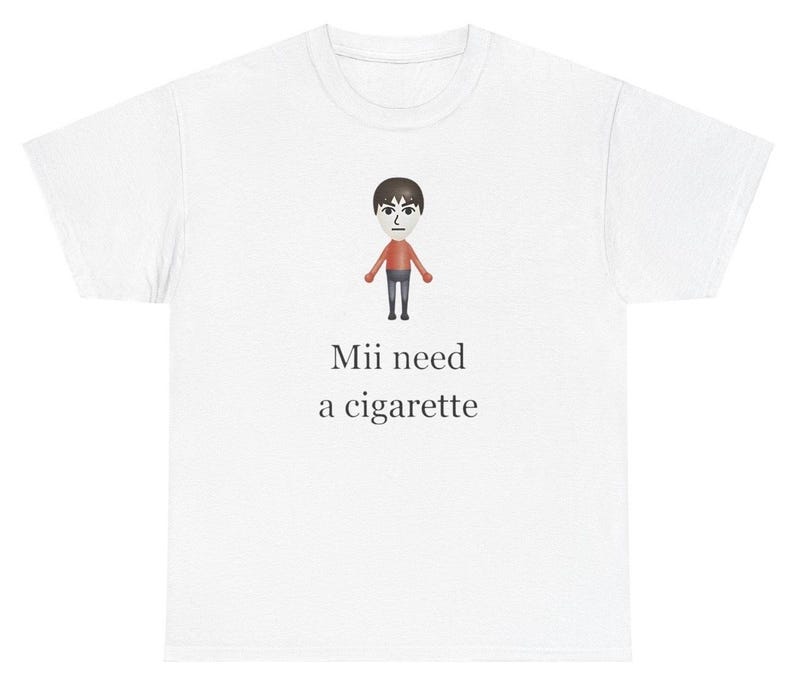 May include: White t-shirt with a cartoon Mii character wearing a red shirt and black pants. The text "Mii need a cigarette" is printed below the character.