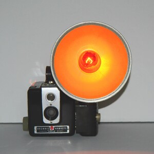 May include: A vintage black Brownie Hawkeye Flash camera with a bright orange flash attachment. The flash is turned on and illuminating the room.