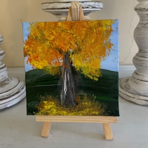 May include: Miniature acrylic painting of a tree with a vibrant yellow and orange canopy, set against a light blue sky and green landscape. The tree trunk is gray and white. The painting is displayed on a small wooden easel.