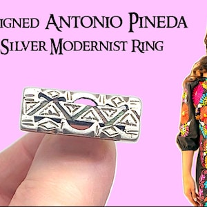 Può includere: Un anello modernista rettangolare in argento 925 con motivi geometrici. L'anello è tenuto davanti a uno sfondo rosa. Il testo "Unsigned Antonio Pineda 925 Silver Modernist Ring" è in alto.