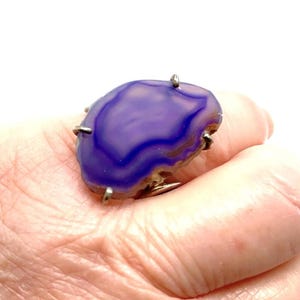 May include: A purple agate ring with a silver setting. The ring is set with a large, irregular shaped agate stone with a swirling pattern of purple and white.