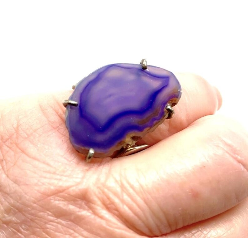 May include: A purple agate ring with a silver setting. The ring is set with a large, irregular shaped agate stone with a swirling pattern of purple and white.