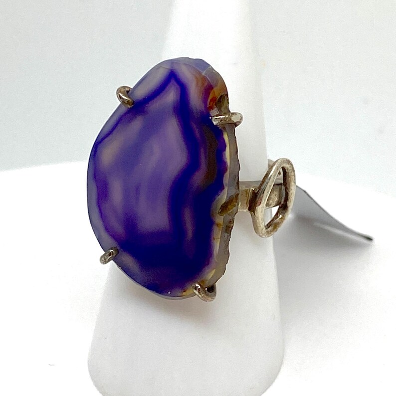 May include: A silver ring with a large, purple agate stone set in the center. The stone has a unique, swirling pattern and is surrounded by silver prongs.