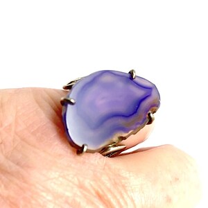 May include: A silver ring with a large, purple agate stone set in the center. The stone has a natural, irregular shape and features a swirling pattern of light and dark purple.