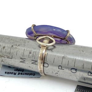 May include: A purple agate gemstone ring with a gold wire setting. The ring is on a metal ruler with markings for sizing.