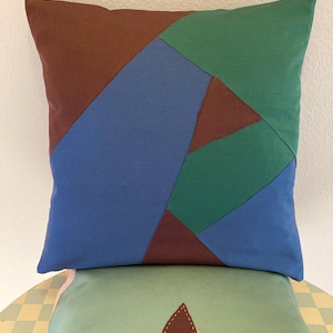 May include: Two decorative throw pillows with geometric patterns. The top pillow is made of blue, green, and brown fabric. The bottom pillow is made of green fabric with a brown leaf design.