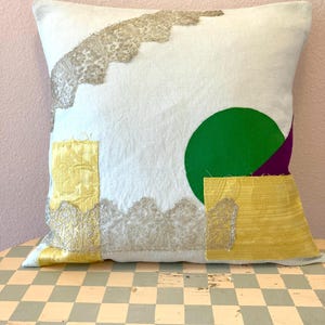 May include: Square white pillow with abstract design. Features a green circle, purple triangle, and gold and silver lace-like embellishments. The pillow is on a checkered surface.