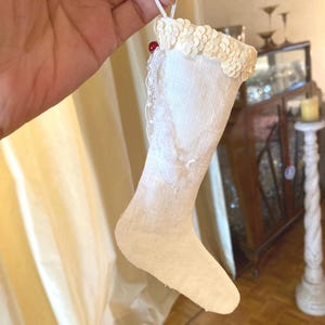 Peut inclure: Une petite chaussette décorative en tissu blanc cassé avec un revers orné de paillettes scintillantes. La chaussette est décorée d'une délicate bordure en dentelle blanche sur le côté et comporte une petite perle rouge près de l'attache.